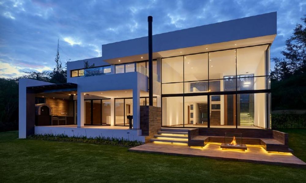 Modern two-story home with large glass windows, illuminated steps, and a cozy outdoor fire pit area, set against a twilight sky.