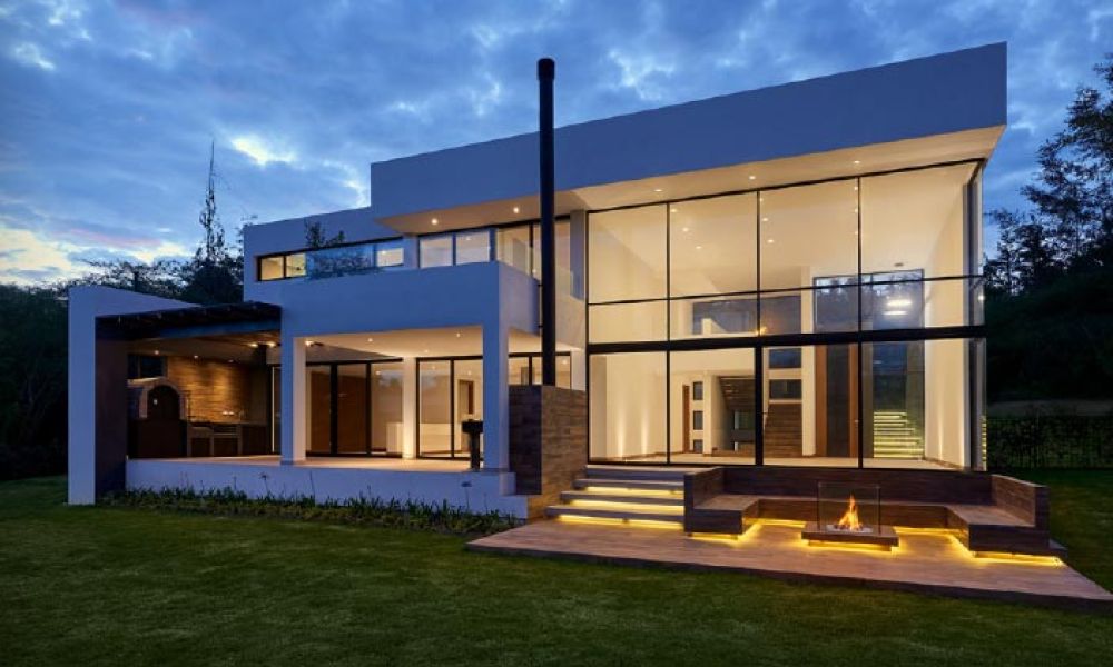 Modern two-story home with large glass windows, illuminated steps, and a cozy outdoor fire pit area, set against a twilight sky.