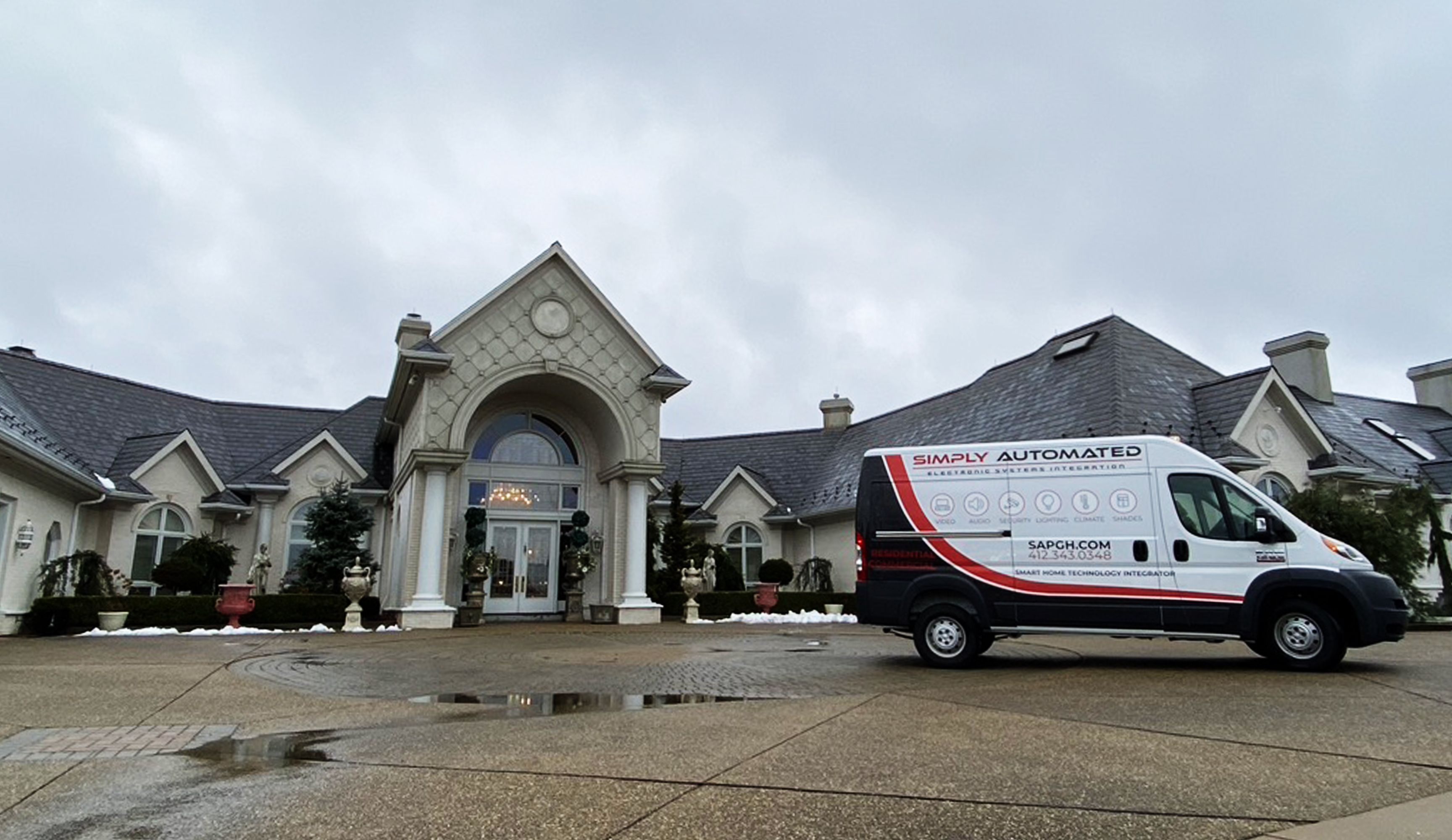 A grand residential property with a large arched entrance, elegant landscaping, and a decorative driveway. A service van is parked in front, highlighting home technology integration services, set against a cloudy sky.