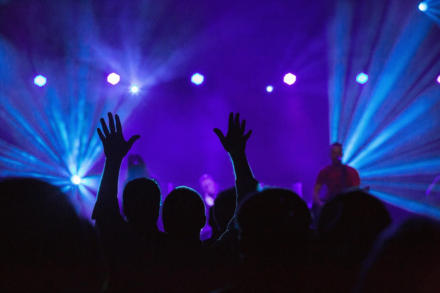 Silhouetted worshippers with raised hands facing a brightly lit stage, creating an immersive worship atmosphere with purple and blue lighting.