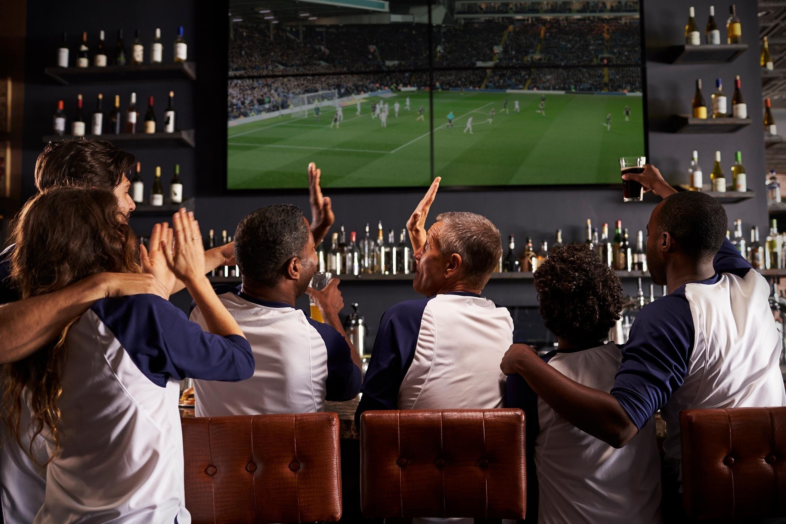 A group of men in a sports bar watching a soccer match in a 4-TV video wall, celebrating a goal.