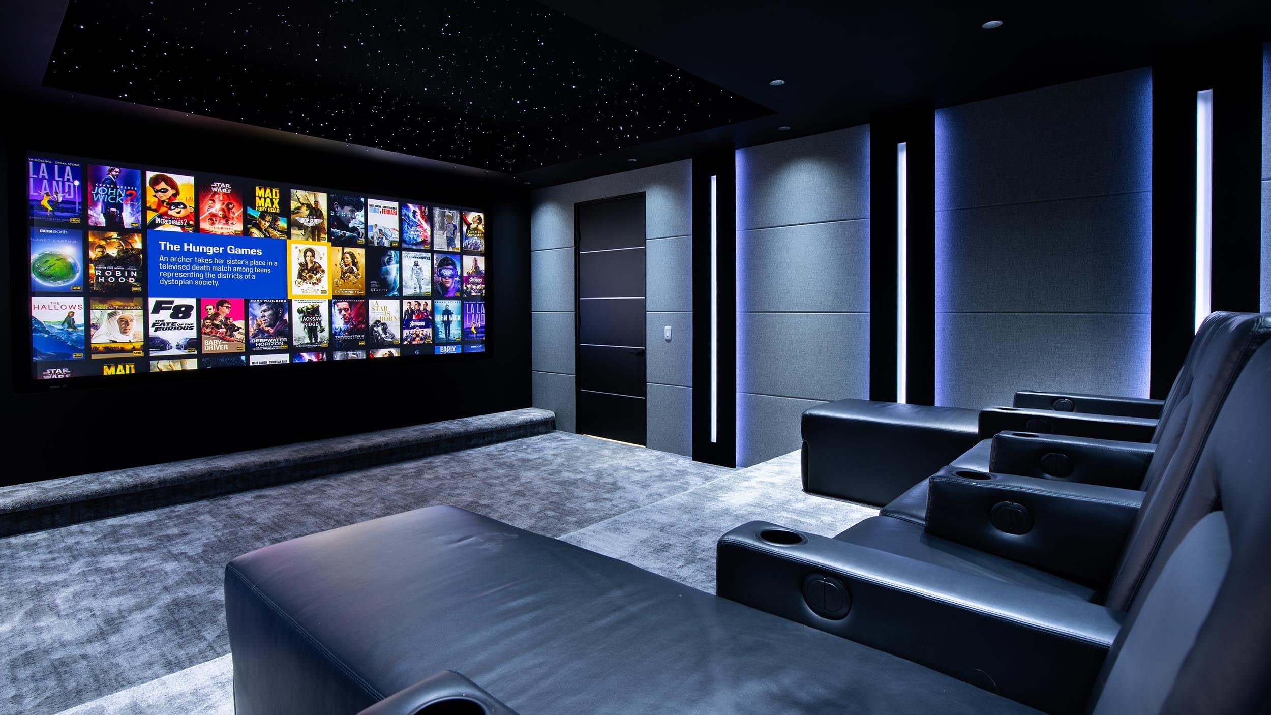 A luxurious home theater featuring plush reclining seats facing a large wall-mounted screen displaying various movie covers. The room is designed with modern acoustic panels, subtle vertical lighting accents, and a ceiling that mimics a starry night sky, creating an immersive cinematic experience.